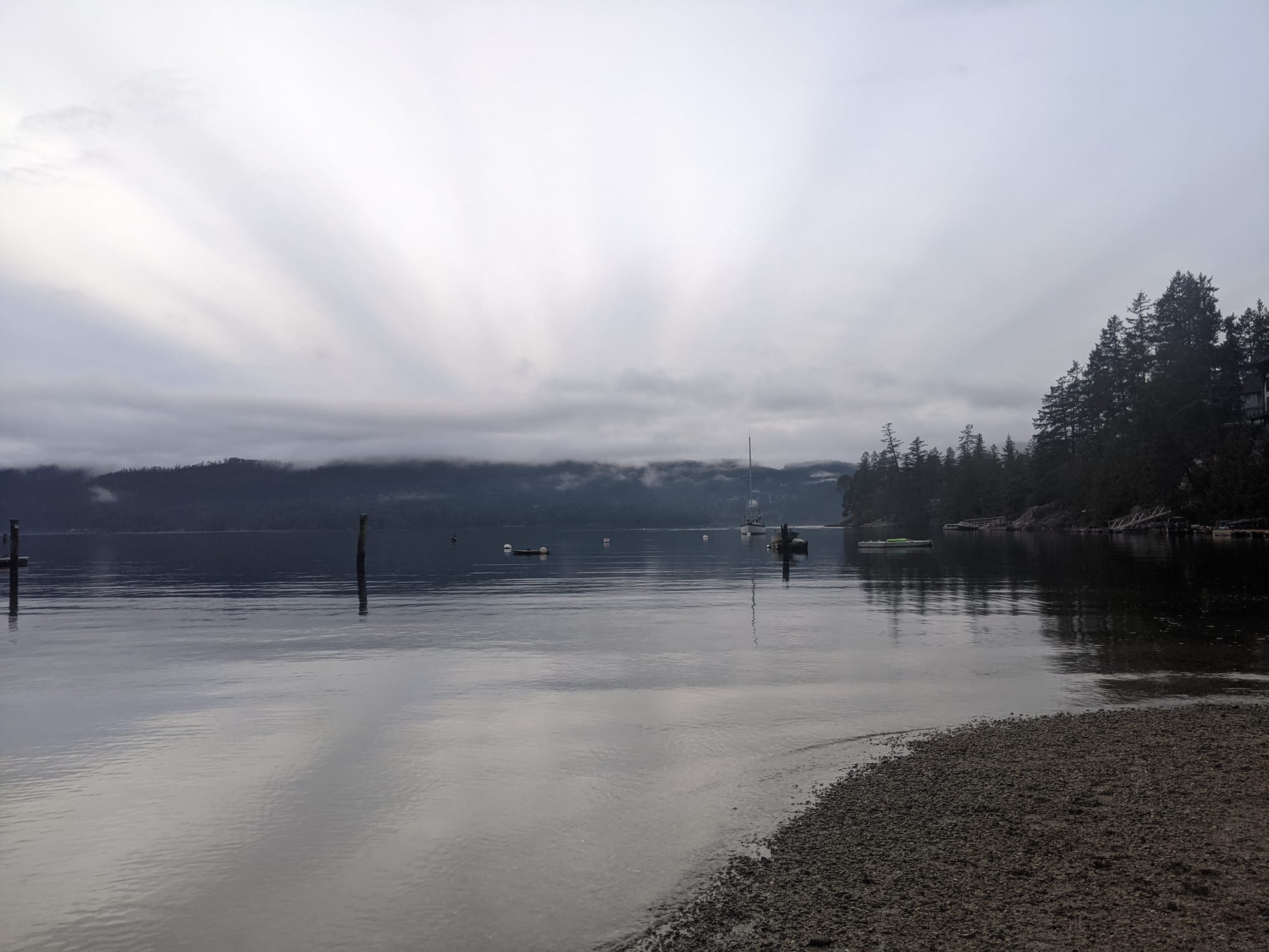 The suns rays scatter in an arc through clouds above fog topped green mountains and an inlet off Salish Sea.