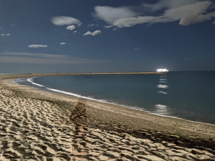 Long shadow of person cast over sands along a coastline with a cruiseship lit in the distance under a full moon sky