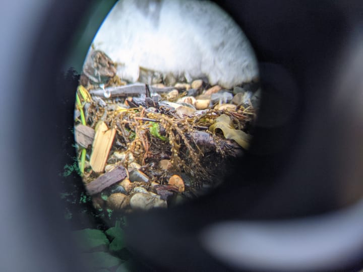 zoomed in oval snapshot of foamy wave crashing against ch'alitch coastline, a greenish band of lens reflection to the left, against grey black backdrop from inside of binoculars' barrel, phot